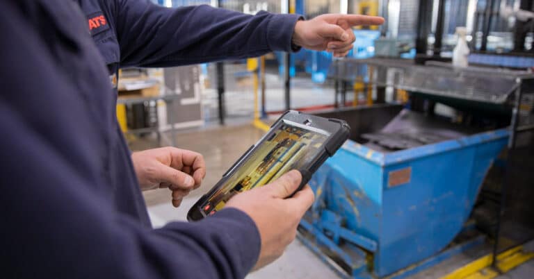 Two technicians on the factory floor holding a tablet with an image of machinery on it.