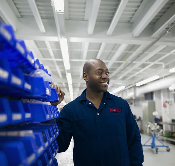 Man in a blue button down looking to the right and touching manufacturing parts.