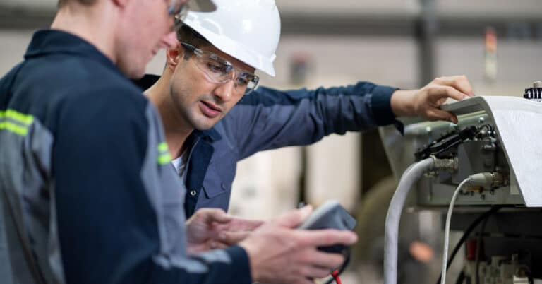 Expert engineer holds a remote control to operate a robotic arm while another engineer checks connections and systems according to programmed commands in a manufacturing plant.