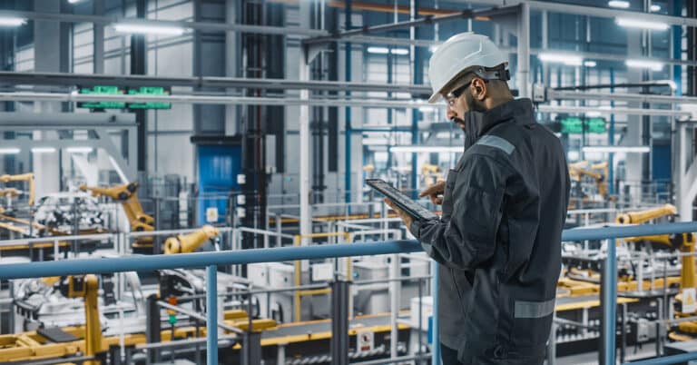 Car factory engineer in work uniform using tablet computer in automotive industrial manufacturing facility.