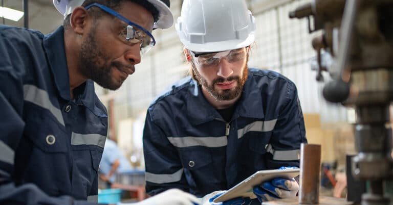 Two male engineers in uniform with helmet using tablet looking drawing design of workpiece material on drilling machine at factory industrial