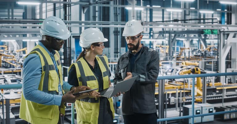 Diverse car factory team collaborates on laptops and tablets, discussing automotive technology in a modern vehicle assembly plant.